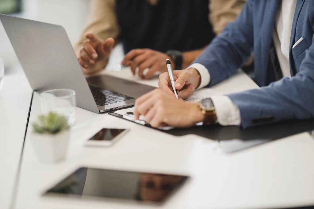 Two colleagues at a bright office desk going through a document on a clipboard, one marking it with a pen beside an open laptop.